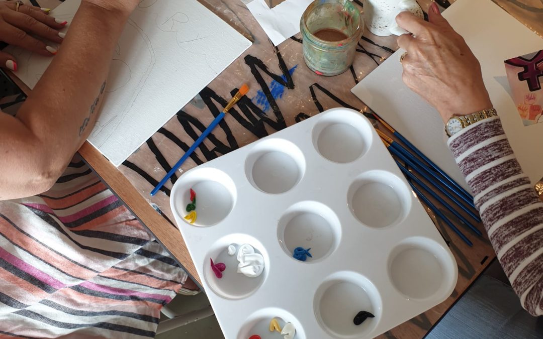 Image of two people with as piece of paper each and paint pot with cups of tea in the background.