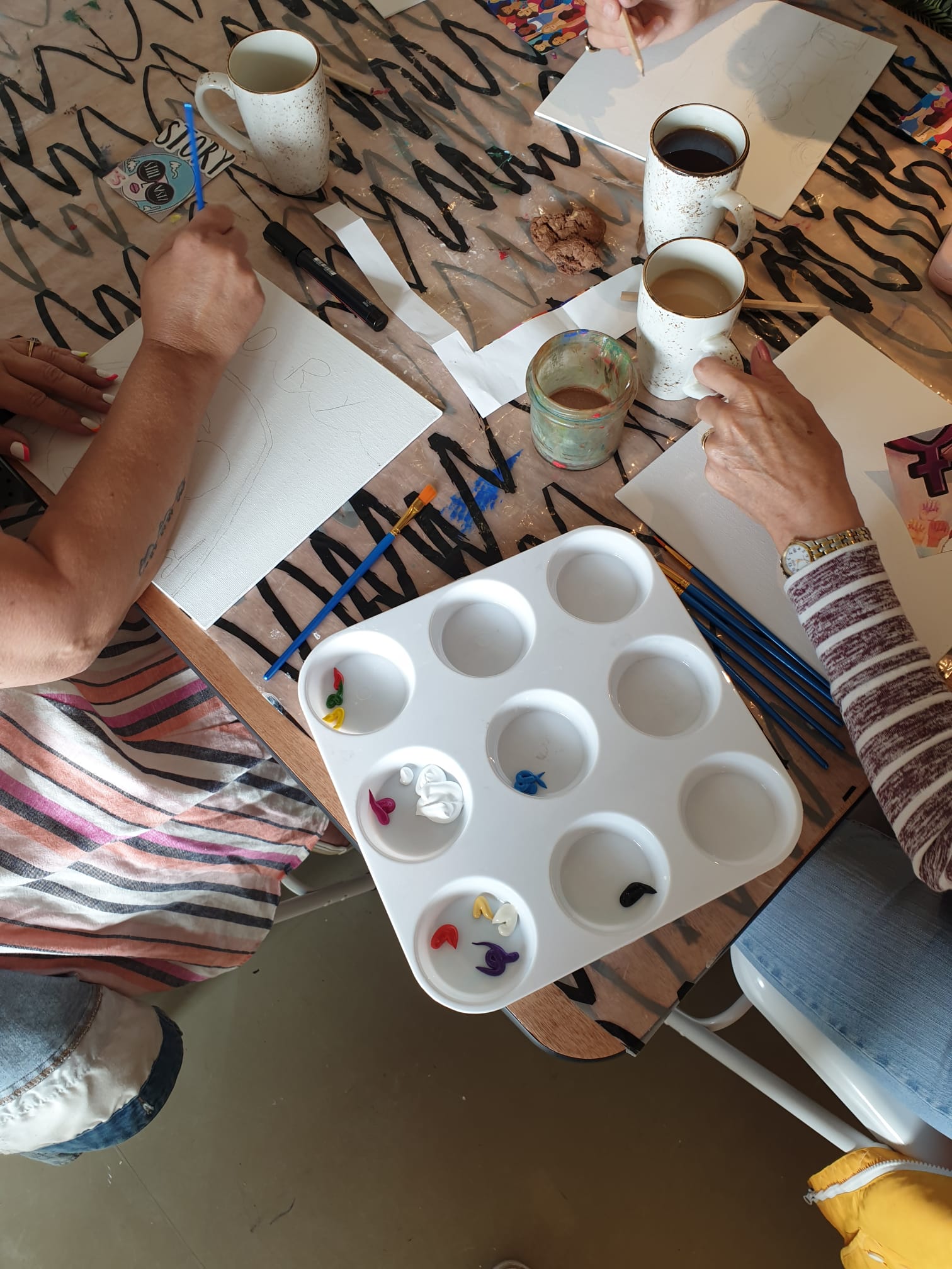 Image of two people with as piece of paper each and paint pot with cups of tea in the background.
