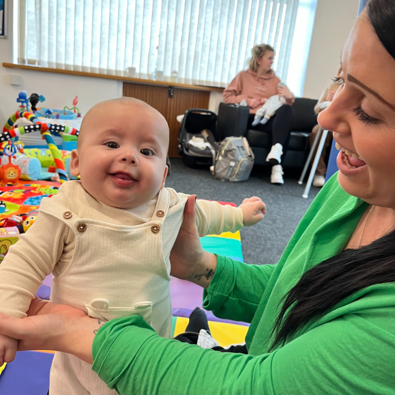 Image shows mum holding her baby both with happy expressions on their faces with another mum and baby in the background