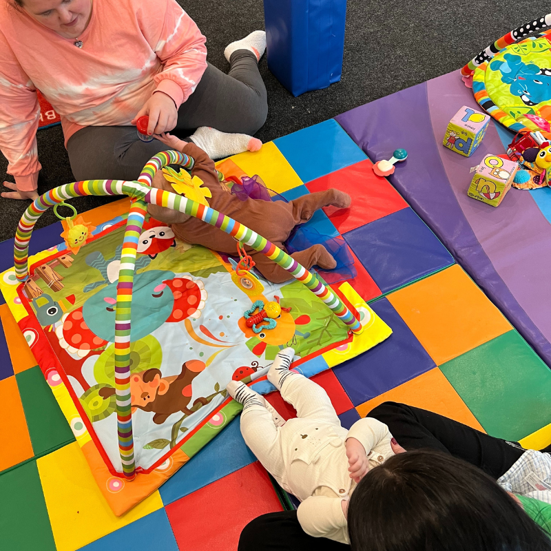Image shows two mums with babies playing on the floormats with some baby toys