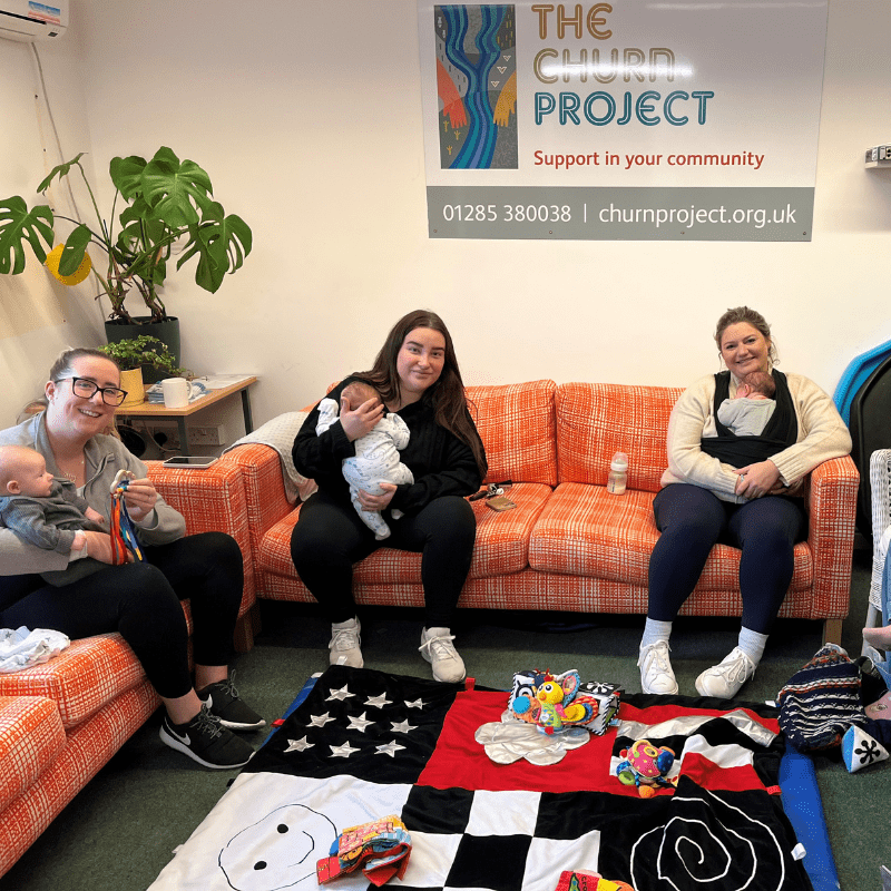 Postnatal Programme Image captures three new mums sitting on the sofas holding their newborn babies smiling at the camera